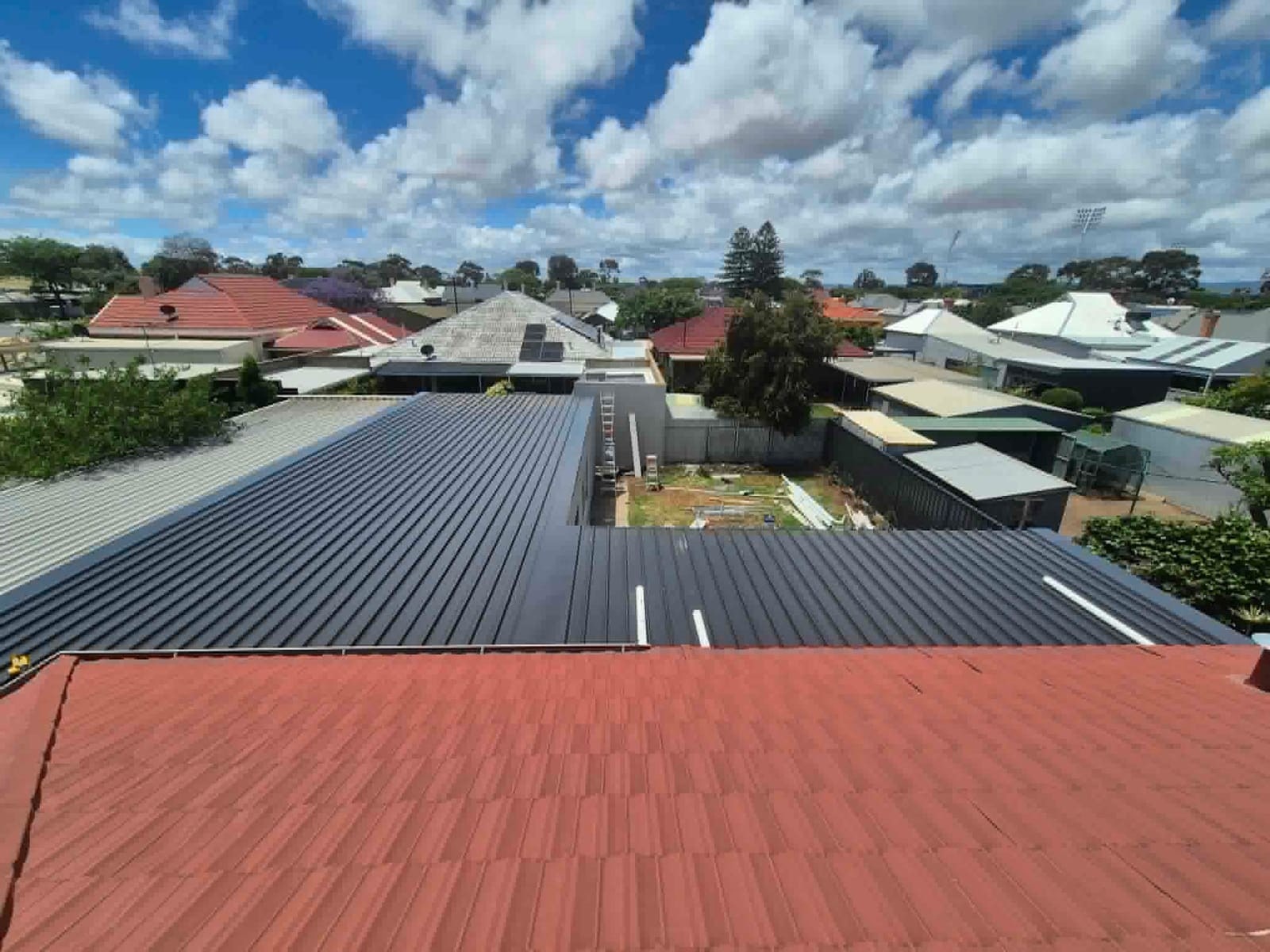 A new Colorbond roof by A1 Roofing, next to an old terracotta tile roof, under a big cloudy sky.