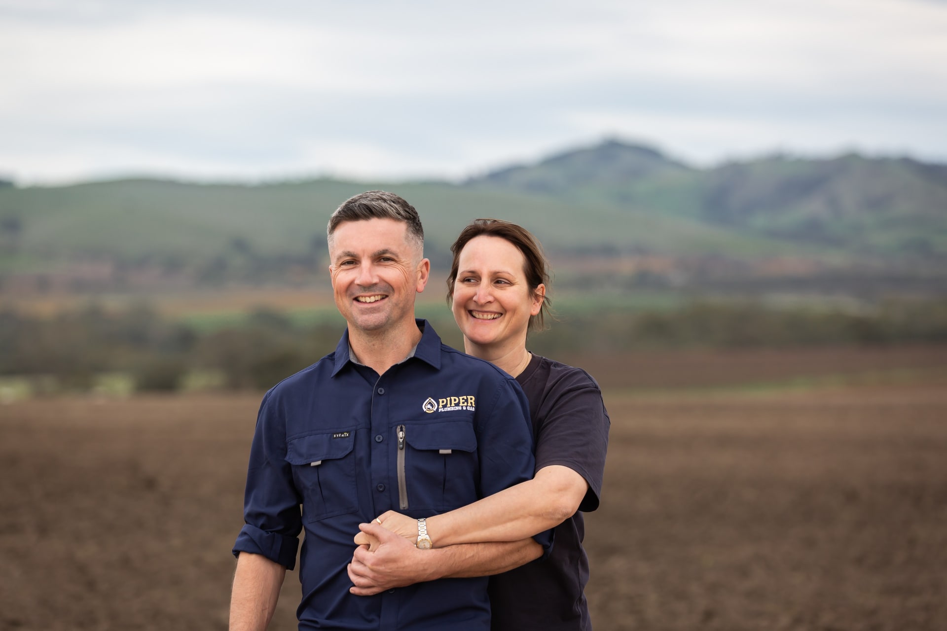 Craig and Hayley of Piper Plumbing & Gas, smiling together in a Barossa paddock with hills behind.