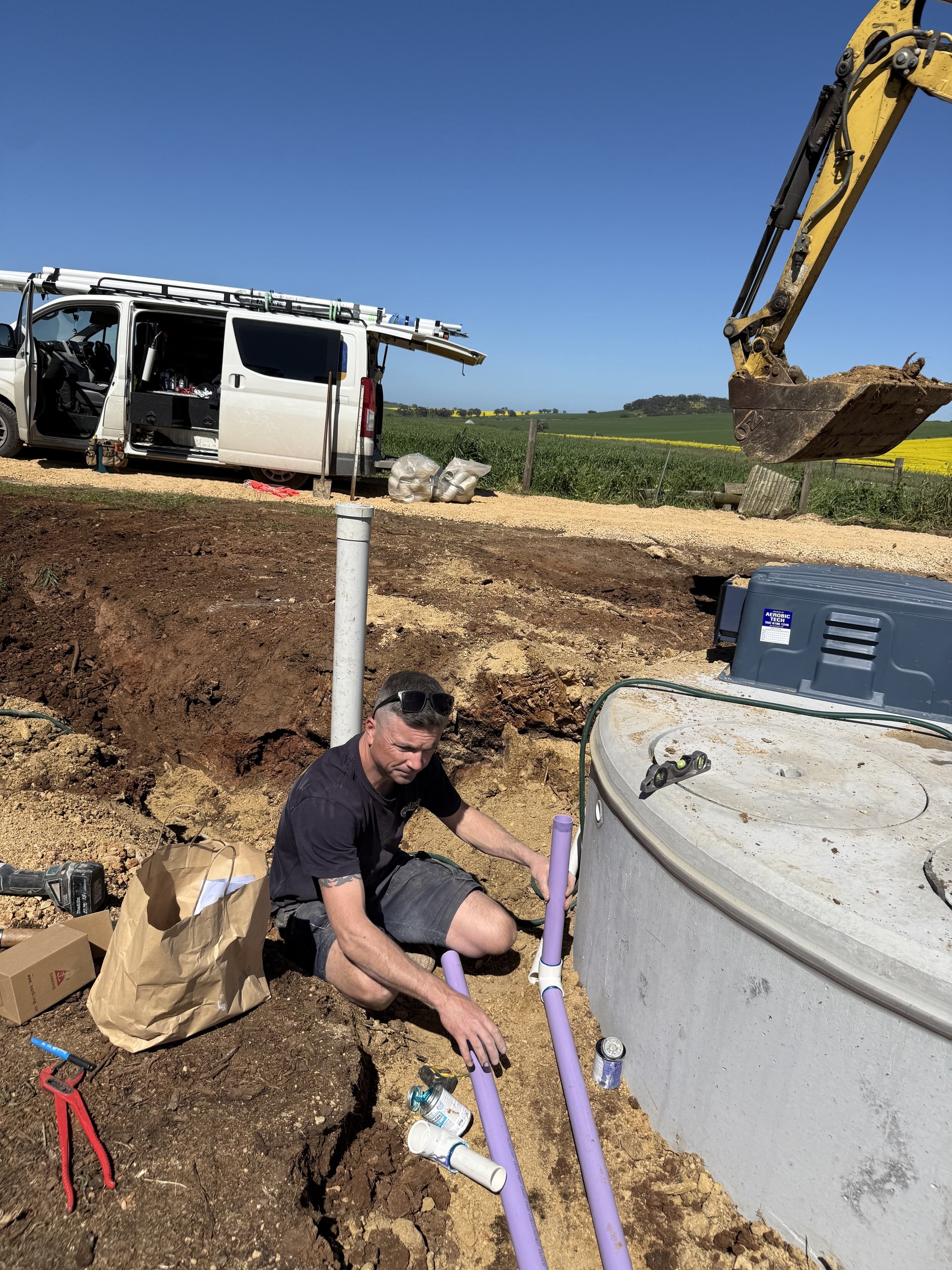 A Piper job site with a tank, the van and an excavator, canola in flower behind.