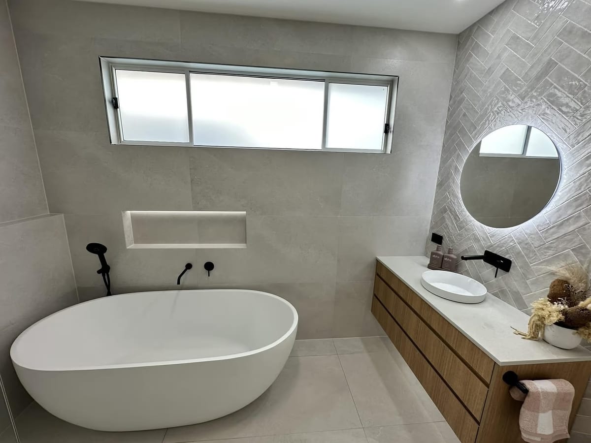 Renovated bathroom with a freestanding bath, wall-hung timber vanity, round backlit mirror and herringbone feature tiles, by ASP Build & Co.