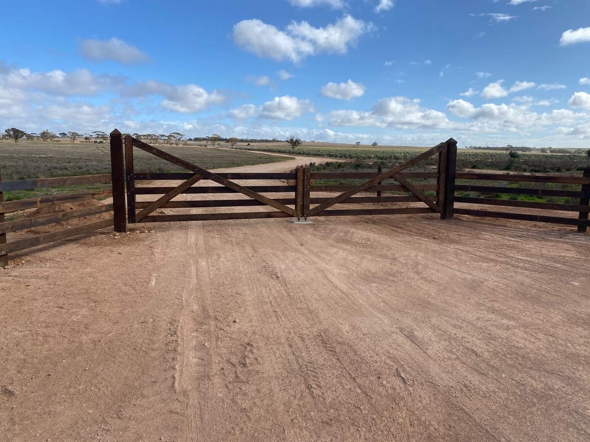 Wide timber farm entrance gates with matching post and rail wings on a dirt driveway, under a big blue sky.