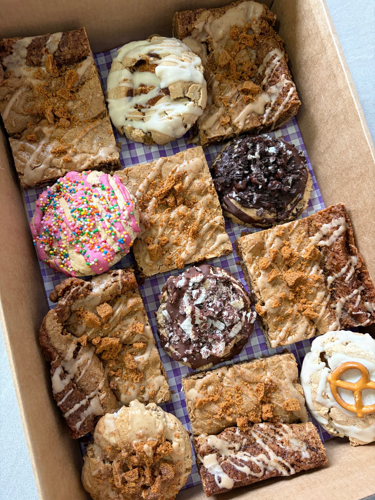 A box of Auggies Kitchen cookies and slices on purple gingham, including a sprinkle-topped cookie and a pretzel-topped cookie.