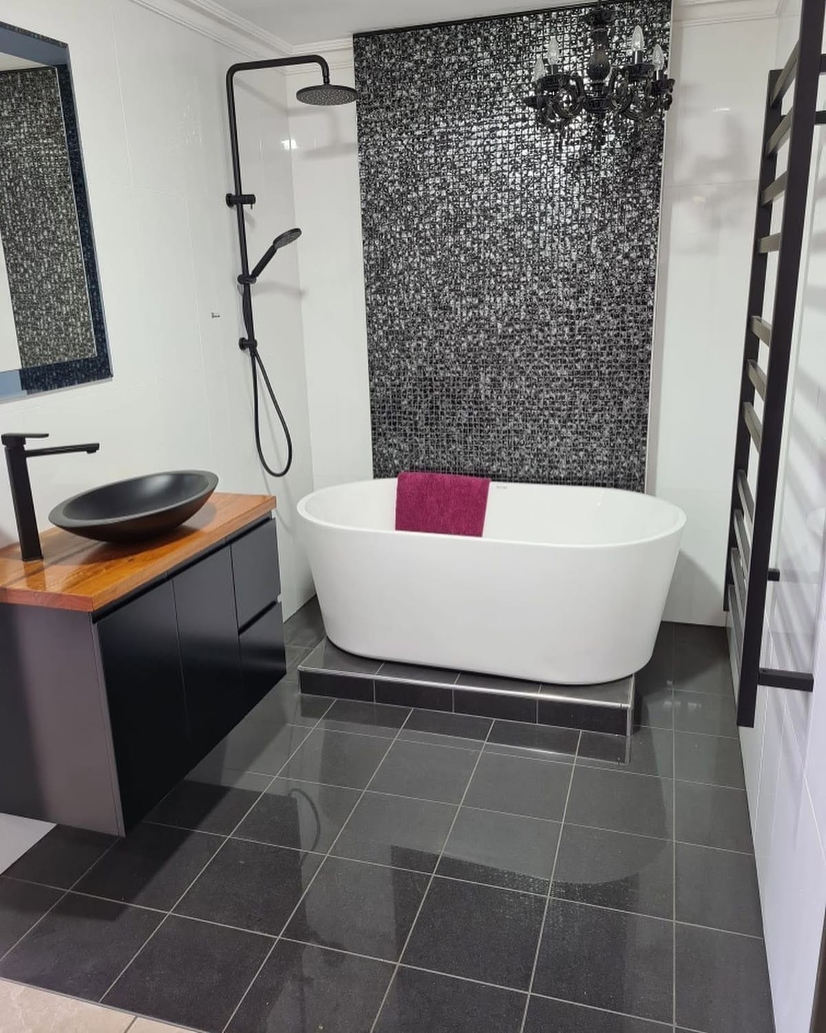 A bathroom with a dark mosaic feature wall behind a freestanding bath, a timber vanity with a black basin and dark floor tiles.