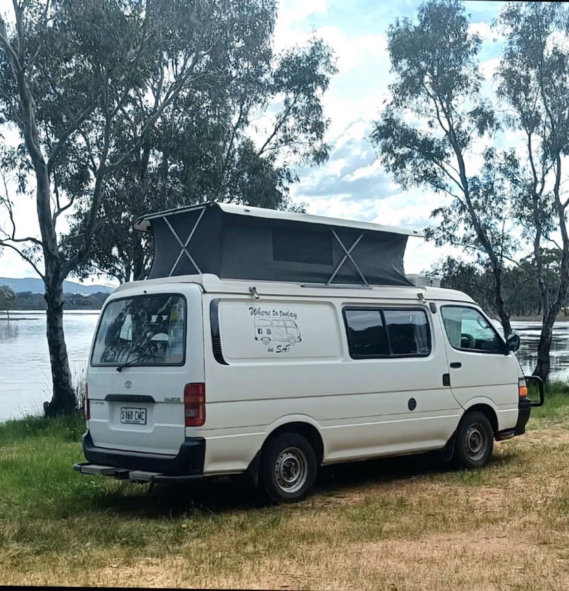Colleen’s white pop-top campervan, Trevor, parked under gum trees beside a lake.