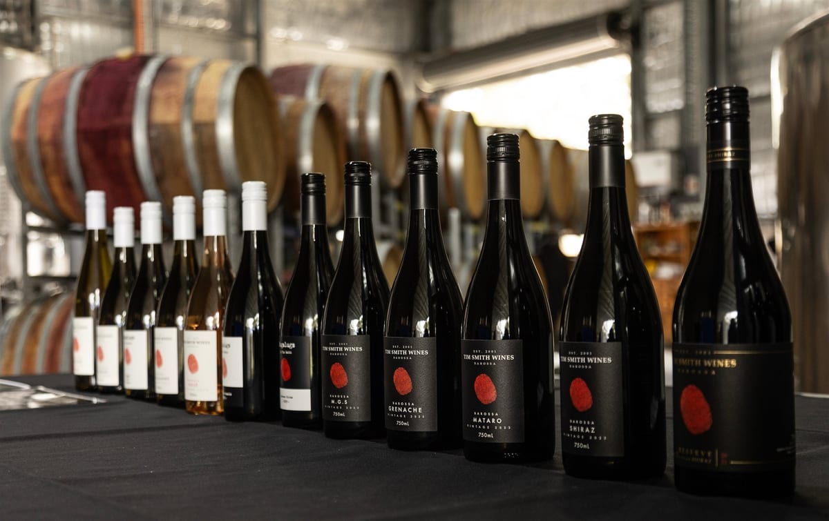 A line of Tim Smith Wines bottles standing in front of oak barrels in a winery shed.