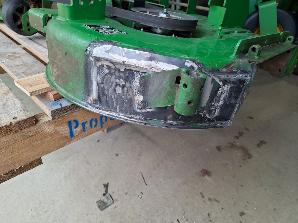 A green ride-on mower cutting deck in a workshop after repair, with new steel plate welded into the front section where it had been torn open.