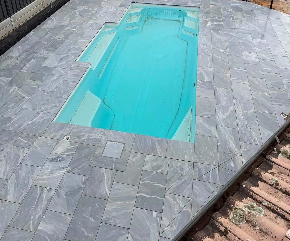 An aerial view of a fibreglass pool wrapped in grey marble-look pavers, with the paving running straight to the water’s edge.