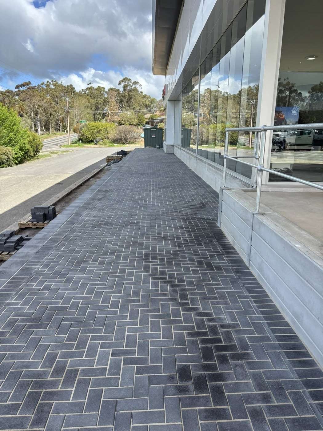 A long commercial walkway beside a car dealership showroom, paved in dark charcoal herringbone brick.