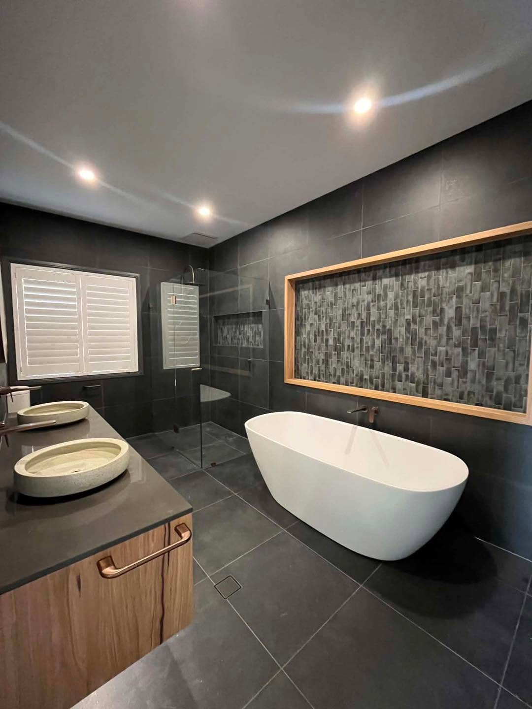 A dark Spriggs & Co master ensuite with a freestanding bath, charcoal tiles, a mosaic feature niche lit in timber, and a double timber vanity.