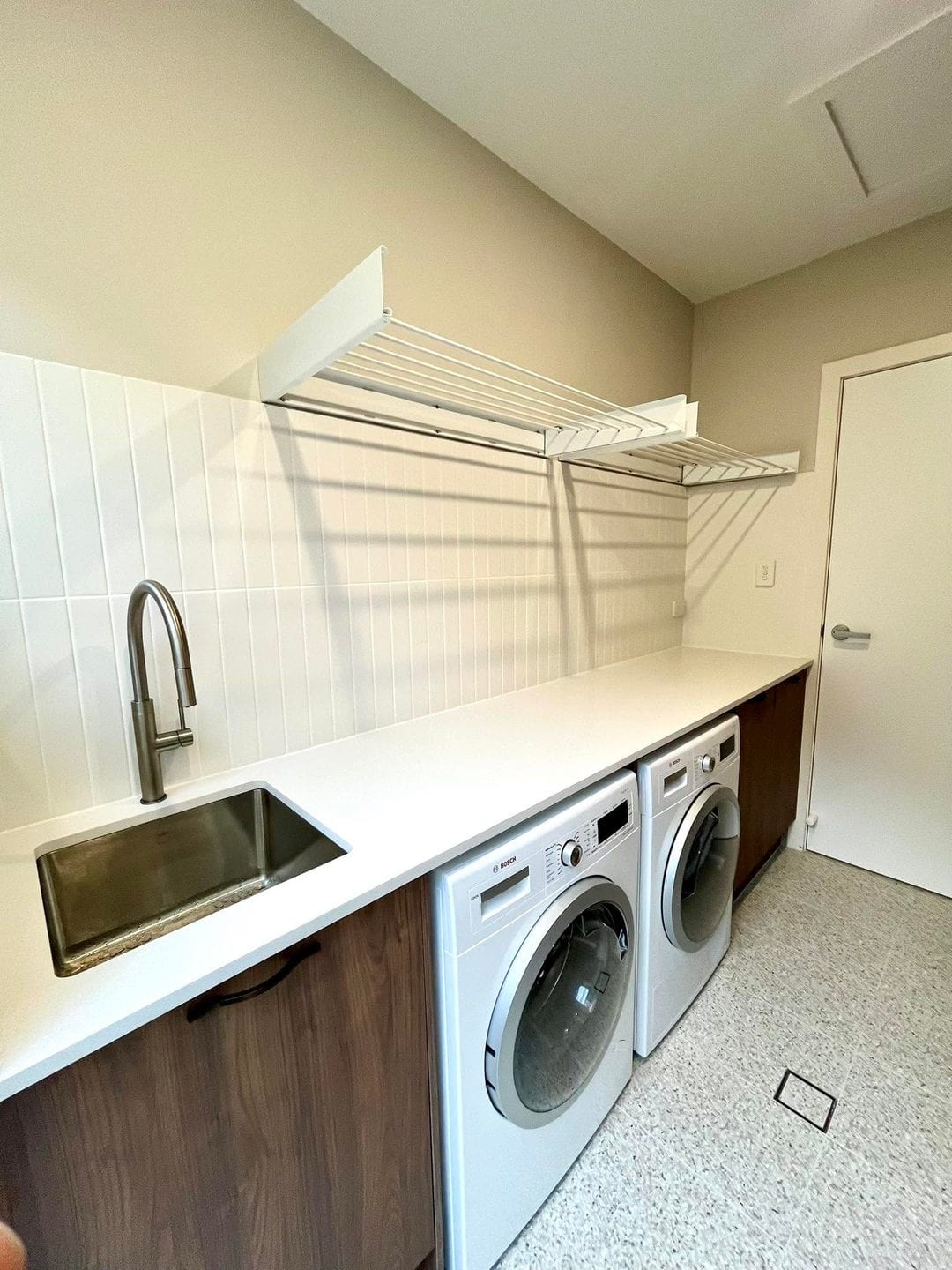 A laundry renovated by Spriggs & Co, with timber-look cabinetry, a white stone benchtop, a stainless sink, two front-loading machines and a drop-down drying rack.