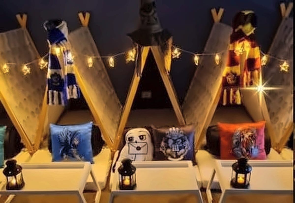 A World of Wizardry teepee setup lit by fairy lights, with house-crest cushions, lanterns and white tray tables.
